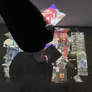 Eine Person steht auf einem Betonboden, auf den Boden und die Wand daneben werden bunte, fragmentierte Bilder projiziert.