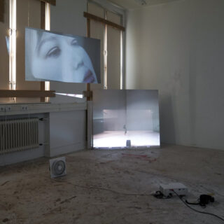 Eine Videoinstallation in einem leeren Raum besteht aus einem großen Bildschirm mit der Nahaufnahme eines Gesichts und einem verspiegelten Bildschirm, auf dem eine kleine menschliche Figur zu sehen ist, mit Kabeln und Geräten auf dem Boden.