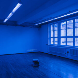 Ein leerer Raum mit Holzboden, großen Fenstern und fluoreszierender Deckenbeleuchtung, beleuchtet von blauem Licht. In der Mitte steht eine kleine Maschine auf dem Boden.