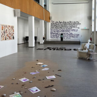 Eine Kunstgalerie mit verstreuten Kunstwerken, Steinen und Sand auf dem Boden, einer Wand mit handgeschriebenem Text, gerahmten Stücken an der Wand und einem Stuhl mit Bezug in der Ecke.