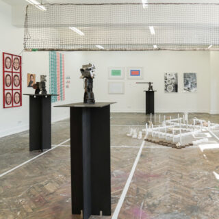 Innenraum einer Kunstgalerie mit Skulpturen auf schwarzen Sockeln, an der Wand montierten Kunstwerken, einer Bodeninstallation aus weißen Stäben und einem von der Decke hängenden Maschennetz.