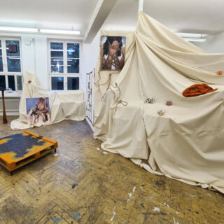 Ein Raum mit drapierten weißen Stoffbespannungen, zwei Fotodrucken einer Person, verstreuten Gegenständen und einer niedrigen Holzplattform auf einem abgenutzten Holzboden.