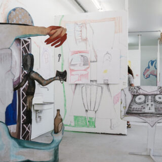 Kunstinstallation in einem Galerieraum mit großen, abstrakten, ausgeschnittenen Figuren, Skizzen auf weißen Wänden und einem DJ-Plattenspieler-Setup aus Pappe.