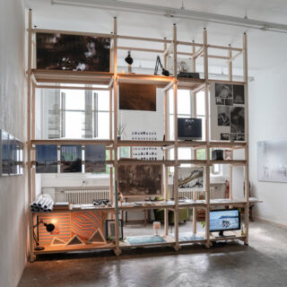 Ein Holzregal voller Fotos, Kunstwerke und anderer Gegenstände steht an einer Fensterwand in einem modernen, minimalistischen Raum. Unten ist ein Schreibtisch mit Monitor und Lampe zu sehen.