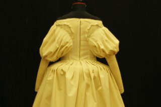 Ein cremefarbenes historisches Kleid mit Puffärmeln und Faltenrock ist auf einer Schneiderpuppe vor schwarzem Hintergrund ausgestellt.