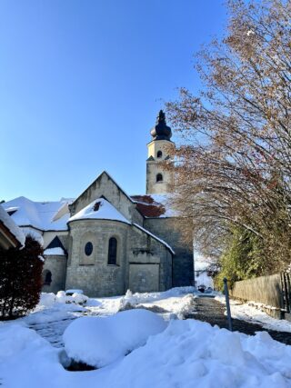 An einem sonnigen Wintertag steht eine Steinkirche mit einem hohen Turm, umgeben von Schnee und kahlen Bäumen unter einem klaren blauen Himmel.
