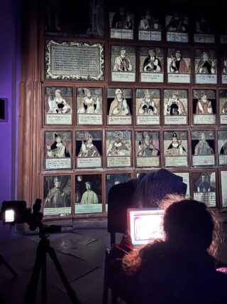 Eine Person sitzt vor einem Laptop und einer Kamera und blickt auf eine beleuchtete Wand mit historischen gemalten Porträts, einige mit beschädigten Gesichtern.
