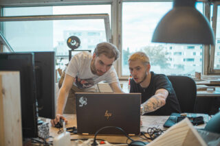 Zwei Personen sitzen an einem Schreibtisch und konzentrieren sich auf einen Laptop-Bildschirm. Im Hintergrund sind Büromaterialien und große Fenster zu sehen.