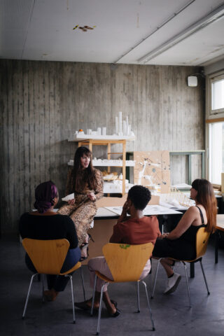 Eine Frau spricht mit drei Personen, die in einem modernen Klassenzimmer im Halbkreis sitzen. Hinter ihr stehen auf Regalen Architekturmodelle.