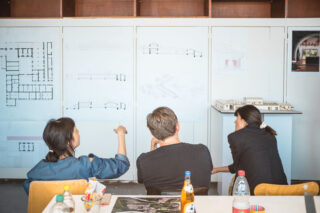 Drei Personen sitzen an einem Tisch und besprechen an einer Wand ausgestellte Architekturpläne. Vor ihnen stehen Getränke und Snacks.