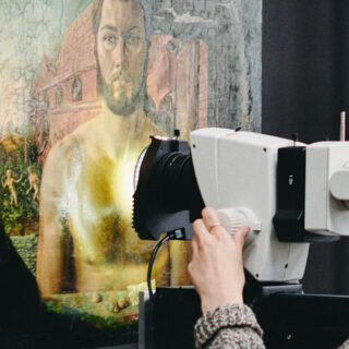 Eine Person bedient ein Scangerät, das auf ein altes, teilweise beschädigtes Gemälde eines bärtigen Mannes mit Ruinen im Hintergrund gerichtet ist.
