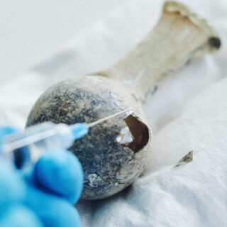 Eine behandschuhte Hand trägt mit einer Spritze Flüssigkeit auf ein gesprungenes, altes Glasgefäß auf, das auf einem weißen Tuch steht.