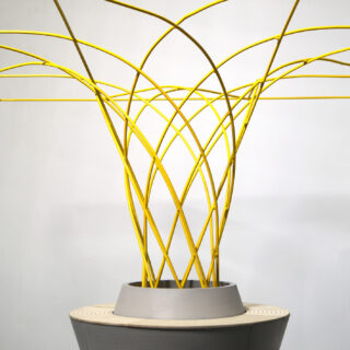 Eine geometrische Skulptur besteht aus sich kreuzenden gelben Stäben, die aus einer grauen, runden Basis hervortreten und vor einem schlichten weißen Hintergrund ein kompliziertes, symmetrisches Muster bilden.