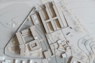 Luftaufnahme eines architektonischen Standortmodells mit Gebäuden, Wegen, kleinen Strukturen und verstreuten Miniaturbäumen auf einer strukturierten topografischen Oberfläche.