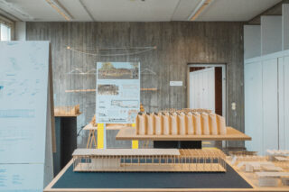 Architekturmodelle und Entwurfstafeln werden auf Tischen und Ständern in einem gut beleuchteten Raum mit Betonwänden und Deckenbeleuchtung ausgestellt.