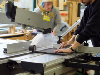 Eine Person bedient eine Tischkreissäge, um ein schwarzes Material zu schneiden, während eine andere Person im Hintergrund in einer Werkstatt arbeitet.