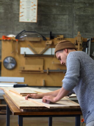 Eine Person mit Mütze und Pullover schleift in einer Holzwerkstatt ein Stück Holz auf einer Werkbank.