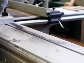 Nahaufnahme einer staubigen Metalloberfläche einer Tischkreissäge mit einem sichtbaren Handabdruck im Staub, der Maschinenhebel und einen unscharfen Hintergrund zeigt.