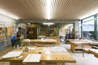 Eine geräumige Holzwerkstatt mit großen Tischen, Werkzeugen und Werkbänken. Eine Person arbeitet an einer Maschine auf der linken Seite des Raumes.