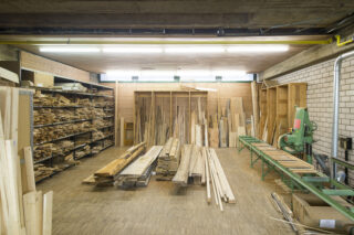 Eine Holzwerkstatt mit Stapeln von Holzbrettern auf Regalen und dem Boden sowie verschiedenen Holzbearbeitungsgeräten an den Wänden.