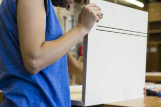 Person in einem blauen ärmellosen Hemd untersucht oder montiert in einer Werkstatt ein Stück helles Holz mit zwei parallelen Rillen.