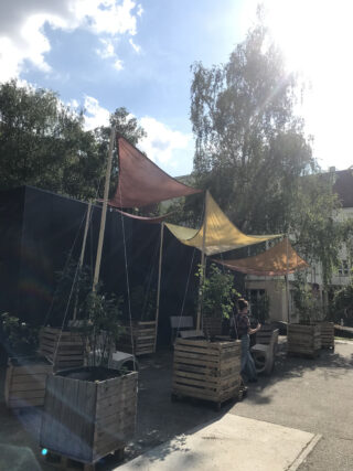 Sitzbereich im Freien mit hölzernen Pflanzgefäßen, bunten Stoffüberdachungen und Menschen, die an einem sonnigen Tag im Schatten sitzen.