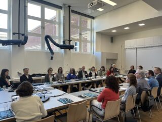 Eine Gruppe von Menschen sitzt in einem hellen Konferenzraum um einen großen U-förmigen Tisch herum und diskutiert, vor ihnen liegen Dokumente und Laptops.