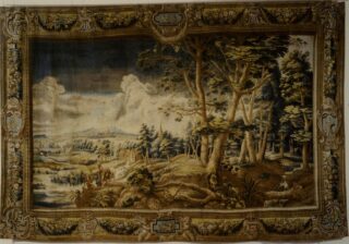 Ein detaillierter Wandteppich zeigt eine Waldlandschaft mit hohen Bäumen, einem Fluss, Hügeln in der Ferne und Figuren am Wasser, alles eingerahmt von einer kunstvollen Bordüre mit dekorativen Motiven.
