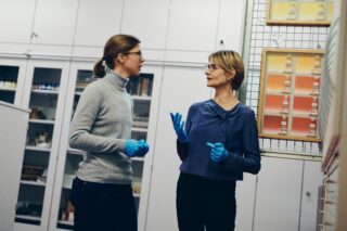 Zwei Personen mit blauen Handschuhen und Brillen unterhalten sich in einer Laborumgebung mit Regalen und wissenschaftlichen Geräten im Hintergrund.