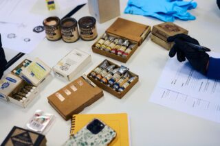 Ein Tisch mit verschiedenen Künstlerutensilien, darunter Farbtuben, Farbdosen, Schachteln, Handschuhe, ein Smartphone, ein Notizbuch und eine behandschuhte Hand, die einen Stift über Papierkram hält.