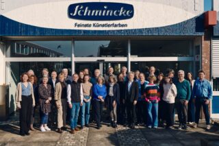Eine Gruppe von Menschen steht vor einem Gebäude mit einem Schild mit der Aufschrift „Schmincke Feine Künstlerfarben“.