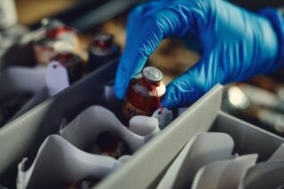 Eine behandschuhte Hand hält ein versiegeltes Fläschchen mit einem Etikett und wählt es aus einer Schachtel mit ähnlichen Fläschchen aus, die durch weiße Trennwände voneinander getrennt sind.