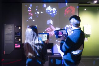 Zwei Personen betrachten eine digitale Ausstellung und halten Tablets in der Hand, auf denen Augmented-Reality-Grafiken angezeigt werden, die zur Projektion an der Wand passen.