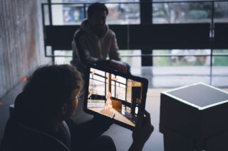 Zwei Personen sitzen drinnen an großen Fenstern; eine hält ein Tablet, auf dem eine Augmented-Reality-Szene mit einer Zeichentrickfigur angezeigt wird.