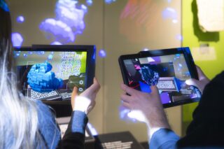 Zwei Personen halten Tablets, auf denen Augmented Reality-3D-Modelle geometrischer Formen und Strukturen angezeigt werden, wobei digitale Grafiken auf die Wände und ihre Hände projiziert werden.