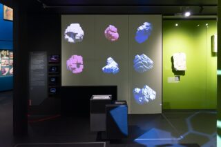 Ausstellungsdisplay mit verschiedenen auf eine Wand projizierten geometrischen Formen, Informationstafeln auf der linken Seite und einer grünen Wand mit einem einzelnen physischen Objekt auf der rechten Seite.