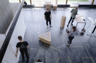 Vier Personen arbeiten an Laptops in einem modernen, offenen Raum mit großen Fenstern und geometrischen Objekten auf dem Boden.