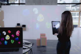 Eine Frau hält einen Laptop vor einer weißen Wand, auf die bunte digitale Formen projiziert werden; ein weiterer Laptop mit ähnlichen Formen steht auf einem Tisch im Vordergrund.