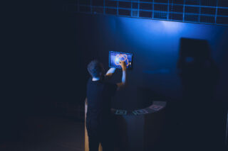 Eine Person interagiert mit einem Touchscreen, der an einer dunkelblauen Wand montiert ist, und zeichnet in einem schwach beleuchteten Raum auf dem Display.