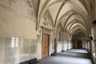 Ein langer, gewölbter Steinflur mit Holztüren, gewölbten Decken und Informationstafeln an einer Wand.