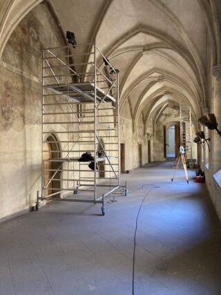 Ein gewölbter Flur mit Gerüsten, Beleuchtungsgeräten und Stativen, aufgestellt für Restaurierungs- oder Filmaufnahmen; links sind verblasste Wandfresken sichtbar.