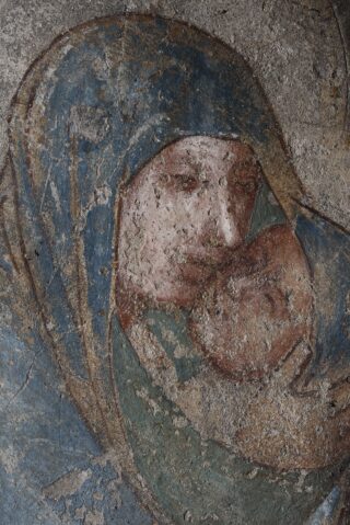 Verblasstes Fresko einer verschleierten Figur, die ein Kind hält; die Oberfläche ist abgenutzt und verwittert, mit sichtbaren Rissen und abgeplatzter Farbe, was auf Alter und Verfall hindeutet.