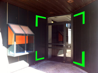 Ein Eingang mit Glastür und schwarzen Paneelen, grünen Eckmarkierungen, einem kleinen Fenster auf der linken Seite und einer Gegensprechanlage an der rechten Wand.