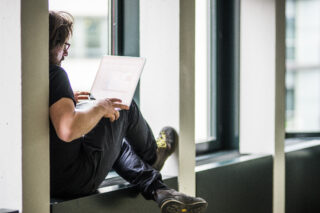 Eine Person sitzt mit gekreuzten Beinen auf einem Fensterbrett und arbeitet an einem Laptop. Im Hintergrund sind große Fenster und eine verschwommene Gebäudefassade zu sehen.