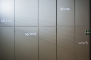 Schränke mit den Worten „Teilen“, „Raum“, „Ideen“ und „Arbeit“ auf den Türen; rechts ist ein kleines grünes Notfallschild zu sehen. Licht scheint diagonal über die Paneele.