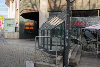 Gläserner Eingangsbau mit unter die Erde führenden Treppen, gelegen auf einem Bürgersteig in der Stadt neben einem Gebäude und einer Restaurantterrasse.