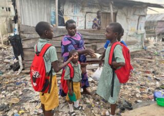 Eine Frau gibt vier Schulkindern in Uniformen und roten Rucksäcken Geld. Sie stehen vor einer Holzkonstruktion in einem Gebiet mit verstreutem Schutt.