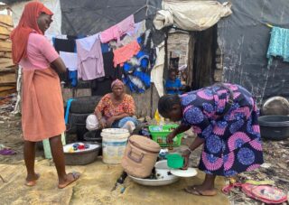 Drei Frauen spülen Geschirr und waschen Wäsche im Freien neben einem provisorischen Unterstand mit hängender Kleidung und verschiedenen Behältern.