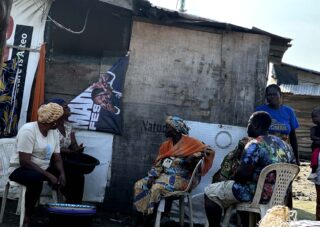 Fünf Personen sitzen und stehen vor einer provisorischen Konstruktion, einige auf Plastikstühlen, und unterhalten sich an einem sonnigen Tag. An der Wand hinter ihnen sind Plakate und Stoffe zu sehen.