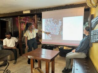 Eine Frau steht da und zeigt auf eine auf eine Leinwand projizierte Karte, während zwei Personen in der Nähe sitzen und ihre Präsentation in einem dekorierten Innenraum verfolgen.